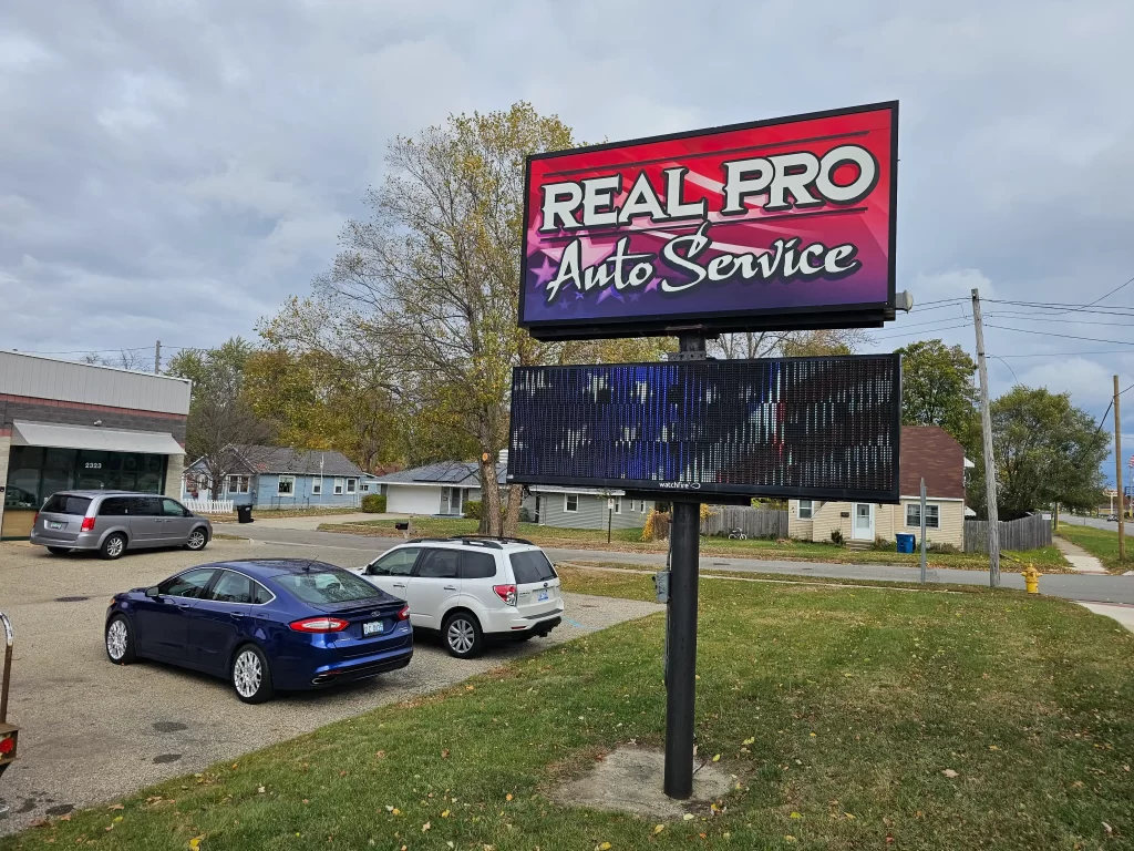 "Real Pro Auto Service" sign with parked cars and houses in the background on a cloudy day.