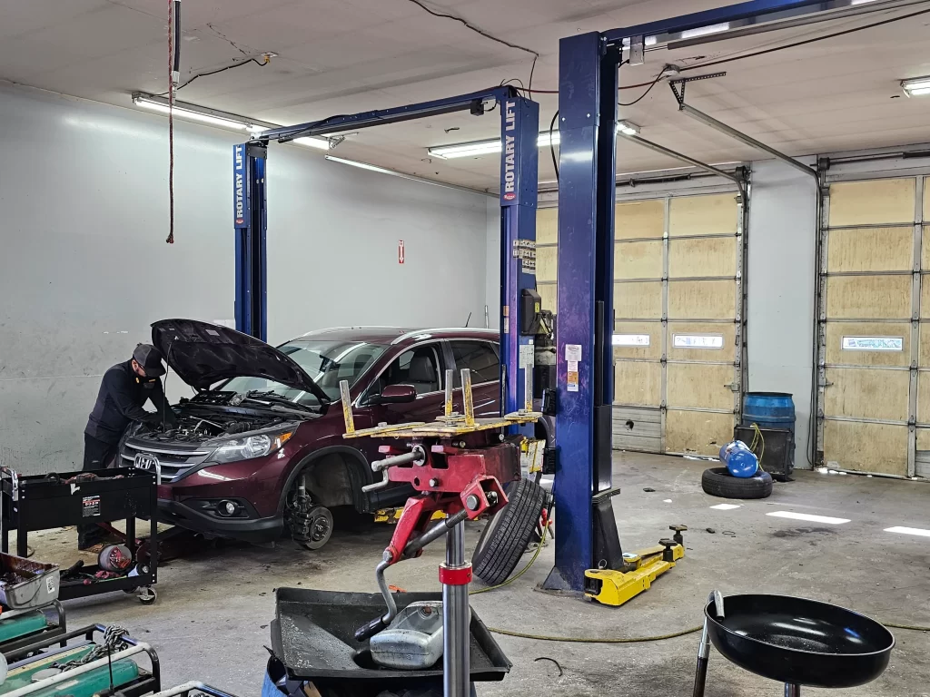 Mechanic working on a maroon SUV with an open hood, inside an auto repair garage with lifts and tools.