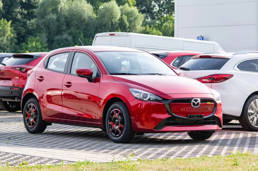 A shiny red Mazda hatchback car parked outdoors, surrounded by other vehicles.