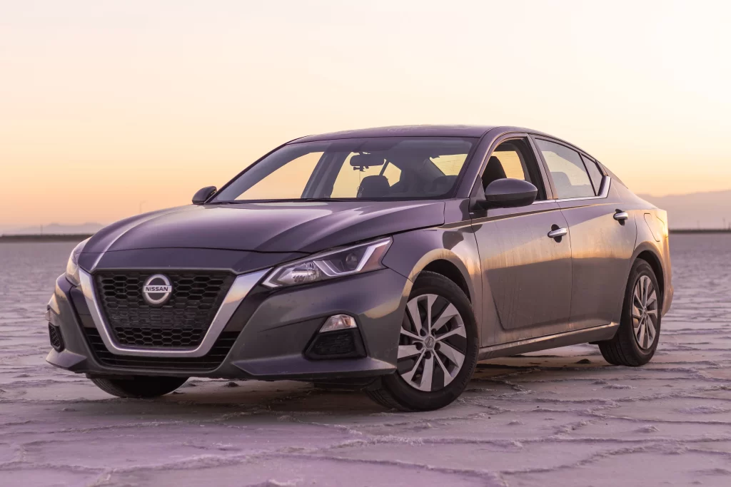 A grey Nissan sedan parked on a flat surface at sunset, showcasing its front and side profile.