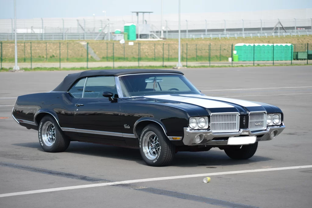 A classic black Oldsmobile convertible with white racing stripes, parked in an empty lot.