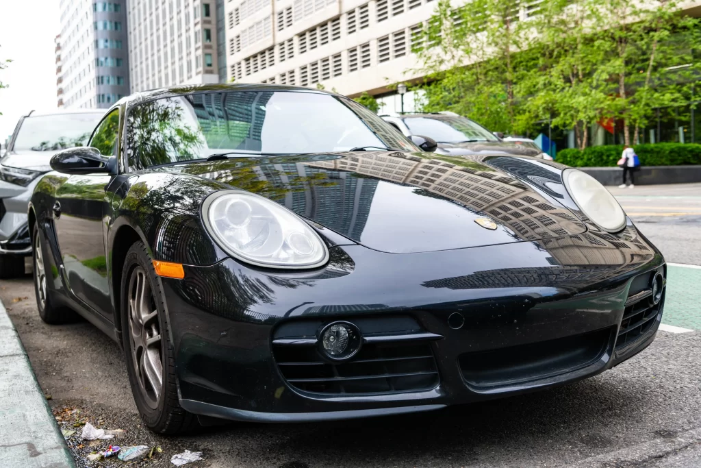 Black Porsche coupe parked on a city street, reflecting buildings and trees in its glossy finish.