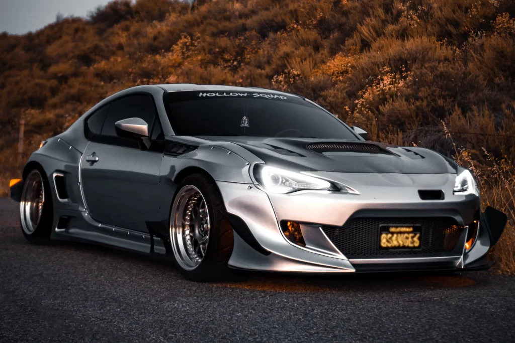 A customized silver Scion coupe with aggressive body modifications parked on a road during sunset.