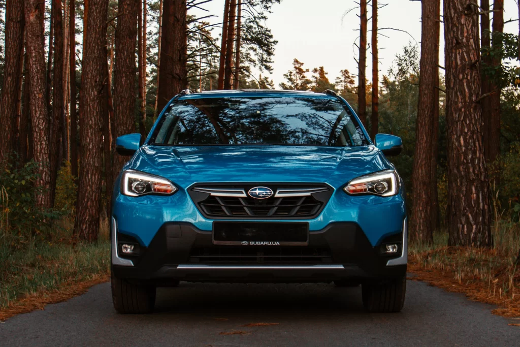 A blue Subaru SUV parked on a forest road, front view with trees in the background.